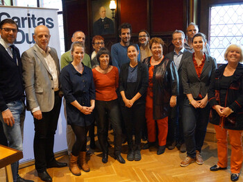 Einige Teilnehmer*innen der Integrationsgespräche der Caritas sind in einem Raum versammeln und posieren vor einem Banner der Caritas.