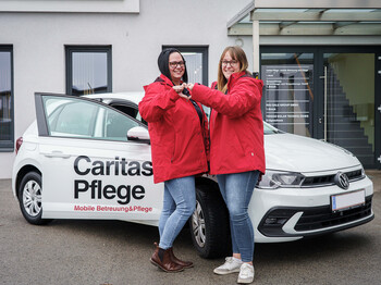 Zwei Caritas Mitarbeiterinnen stehen vor dem Caritas Auto der Mobilen Pflege und formen mit den Händen ein Herz.