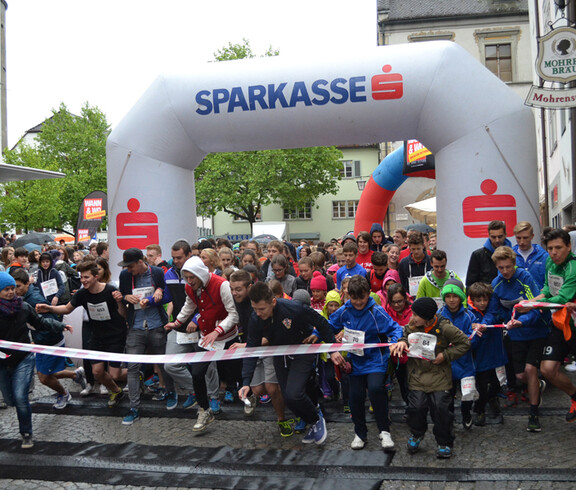 Eine große Ansammlung von Jugendlichen befindet sich in einer Innenstadt. Sie alle nehmen teil an einem Laufwettbewerb. Alle haben sie Wettkampfnummern. 