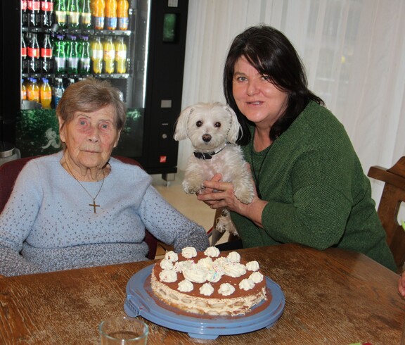 Eine Bewohnerin sitzt am Tisch vor ihrer Geburtstagstorte. Daneben sitzt eine Dame, die einen kleinen weißen Hund im Arm halten, und lächeln gemeinsam in die Kamera.