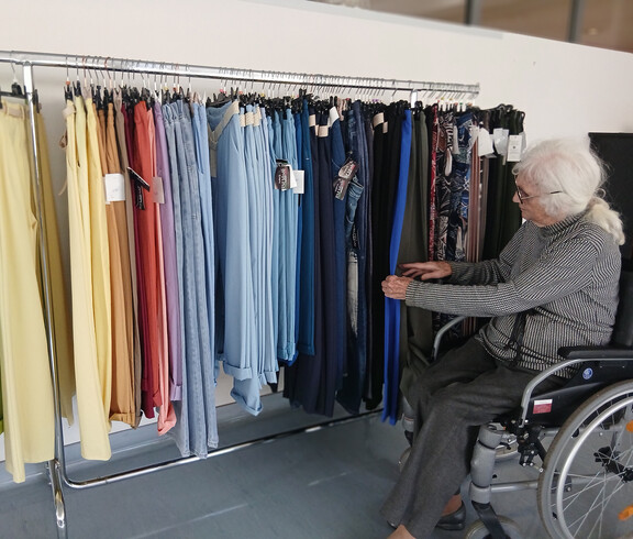 Bewohnerin im Rollstuhl betrachtet Hosen in vielen Farben an einem Kleiderständer