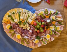 Auf einem großen Holzbrett in herzform ist eine reichhaltige Jausenplatte mit verschiedenen Käsesorten, Wurst, Schinken, gekochten Eiern, Paprikastreifen, Trauben und dekorativ geschnitztem Gemüse angerichtet, die liebevoll mit Kräutern garniert ist.