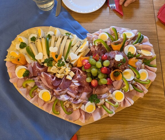 Auf einem großen Holzbrett in herzform ist eine reichhaltige Jausenplatte mit verschiedenen Käsesorten, Wurst, Schinken, gekochten Eiern, Paprikastreifen, Trauben und dekorativ geschnitztem Gemüse angerichtet, die liebevoll mit Kräutern garniert ist.
