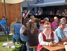 Viele Menschen sitzen an Bierbänken im Garten, im Hintergrund Musiker