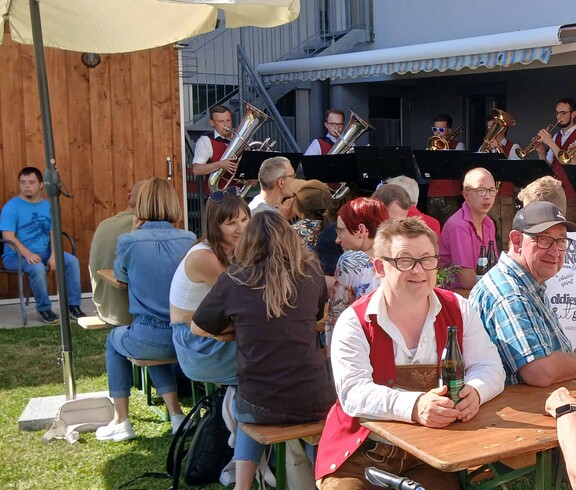 Viele Menschen sitzen an Bierbänken im Garten, im Hintergrund Musiker