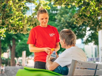 „Eine Person in rotem Caritas-&Du-T-Shirt überreicht einer auf einer Parkbank sitzenden Person Getränke und Hilfsartikel im Freien unter schattenspendenden Bäumen.“
