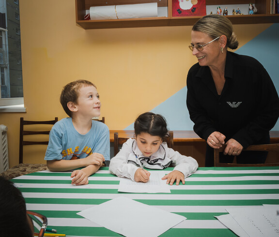 Zwei kleine Kinder grinsen ihre Betreuerin beim Hausaufgaben machen an.
