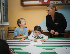 Zwei kleine Kinder grinsen ihre Betreuerin beim Hausaufgaben machen an.