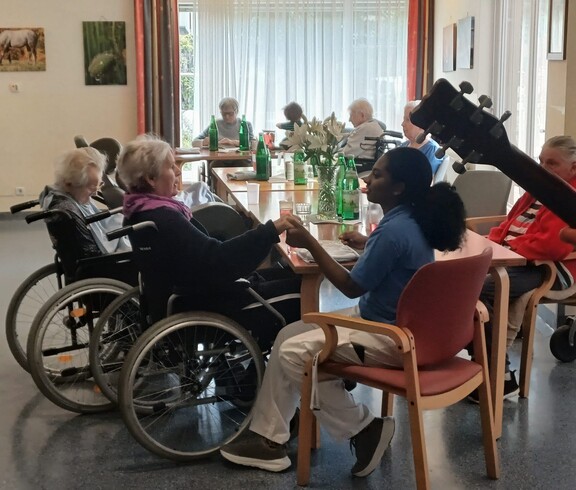Mehrere Bewohnerinnen und Bewohner sitzen gemeinsam mit einer Betreuungsperson an Tischen, halten sich teilweise an den Händen und verbringen bei Musik einen geselligen Moment im Aufenthaltsraum.