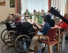 Mehrere Bewohnerinnen und Bewohner sitzen gemeinsam mit einer Betreuungsperson an Tischen, halten sich teilweise an den Händen und verbringen bei Musik einen geselligen Moment im Aufenthaltsraum.