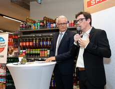 Zwei Personen sprechen mit Mikrofon vor SPAR-Logo, stehen vor Getränkeregal im Markt.