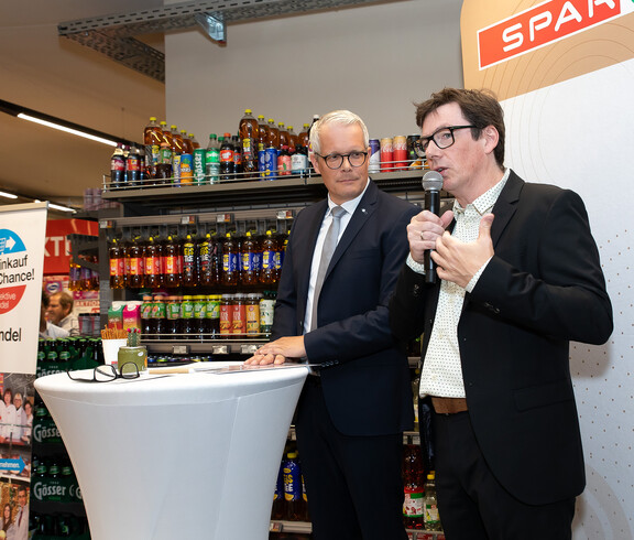 Zwei Personen sprechen mit Mikrofon vor SPAR-Logo, stehen vor Getränkeregal im Markt.