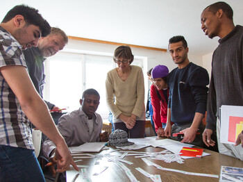 Flüchtlinge aus Afrika und dem nahen Osten lernen Deutsch. Sie sitzen gespannt und konzentriert an einem Tisch und blicken auf ihre Unterlagen. Eine Lehrerin hilft ihnen dabei den Stoff besser aufzunehmen und steht für jede Frage offen. 
