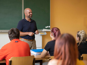 Ein Lehrer steht vorne an der Tafel, die Schüler*innen sehen aufmerksam zu ihm.