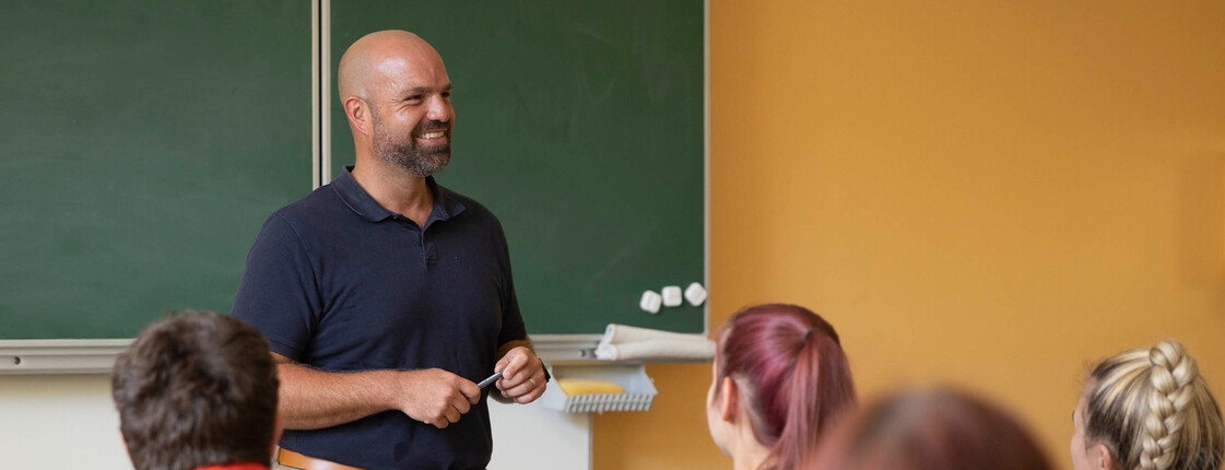 Ein Lehrer steht vorne an der Tafel, die Schüler*innen sehen aufmerksam zu ihm.