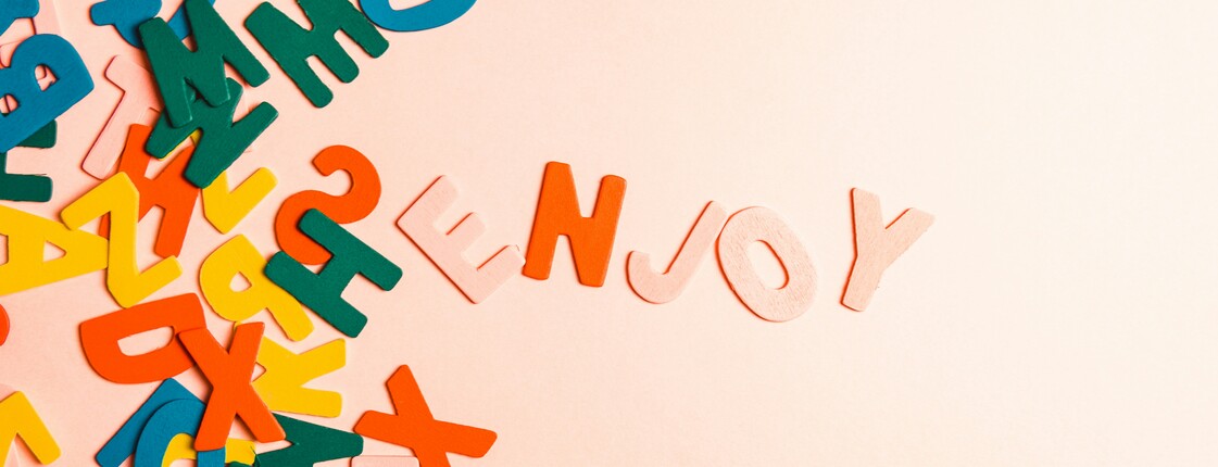 Bunte Holzbuchstaben liegen vertreut auf einem hellen Hintergrund und ein paar Buchstaben bilden das Wort 'enjoy'