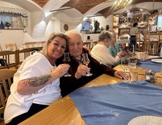 Ein Bewohner und eine Dame der Frauenbewegung sitzen herzlich nebeneinander an einem gedeckten Tisch und halten gemeinsam kleine Gläser zum Anstoßen hoch.
