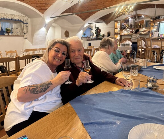 Ein Bewohner und eine Dame der Frauenbewegung sitzen herzlich nebeneinander an einem gedeckten Tisch und halten gemeinsam kleine Gläser zum Anstoßen hoch.