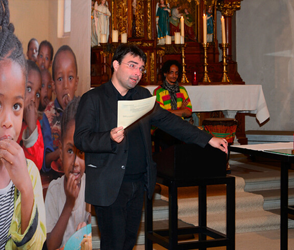 Ein Herr mittleren Alters mit gänzlich schwarzer Kleidung steht am Rednerpult der frastanzer Pfarrkirche. Der Hintergrund ziert eine großes Plakat von afrikanischen Kindern und einem afrikanischen Trommelspieler. 