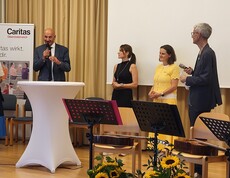 Fünf Personen stehen auf einer Bühne. Ein Mann spricht ins Mikrofon.
