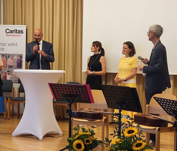 Fünf Personen stehen auf einer Bühne. Ein Mann spricht ins Mikrofon.