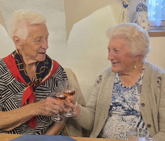 Zwei Bewohnerinnen sitzen nebeneinander an einem Tisch, halten jeweils ein kleines Glas in der Hand und stoßen miteinander an, während vor ihnen Geschirr und Getränke auf dem Tisch stehen.