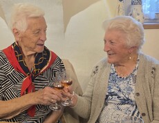 Zwei Bewohnerinnen sitzen nebeneinander an einem Tisch, halten jeweils ein kleines Glas in der Hand und stoßen miteinander an, während vor ihnen Geschirr und Getränke auf dem Tisch stehen.