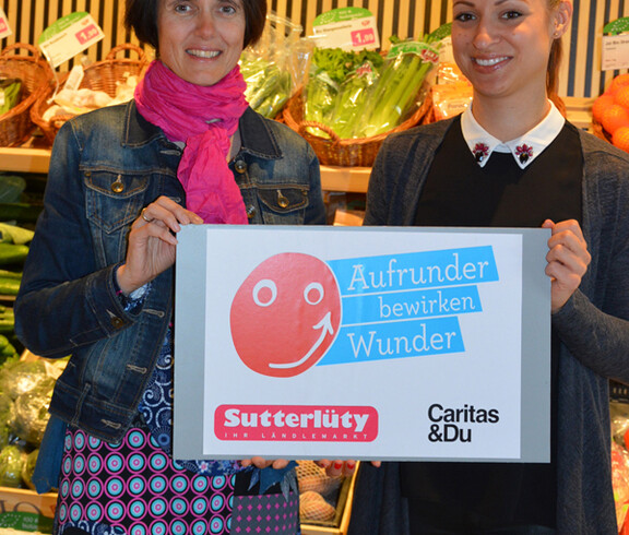 Caritas-Mitarbeiterin Doris Jenny stellt mit einer Sutterlüty-Angestellten die Aktion "Aufrunden" vor. Sie halten dabei ein Schild in der Hand, das die Aktion illustirert, Im Hintergrund befindet sich ein Regal mit Lebensmitteln. 