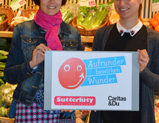 Caritas-Mitarbeiterin Doris Jenny stellt mit einer Sutterlüty-Angestellten die Aktion "Aufrunden" vor. Sie halten dabei ein Schild in der Hand, das die Aktion illustirert, Im Hintergrund befindet sich ein Regal mit Lebensmitteln. 