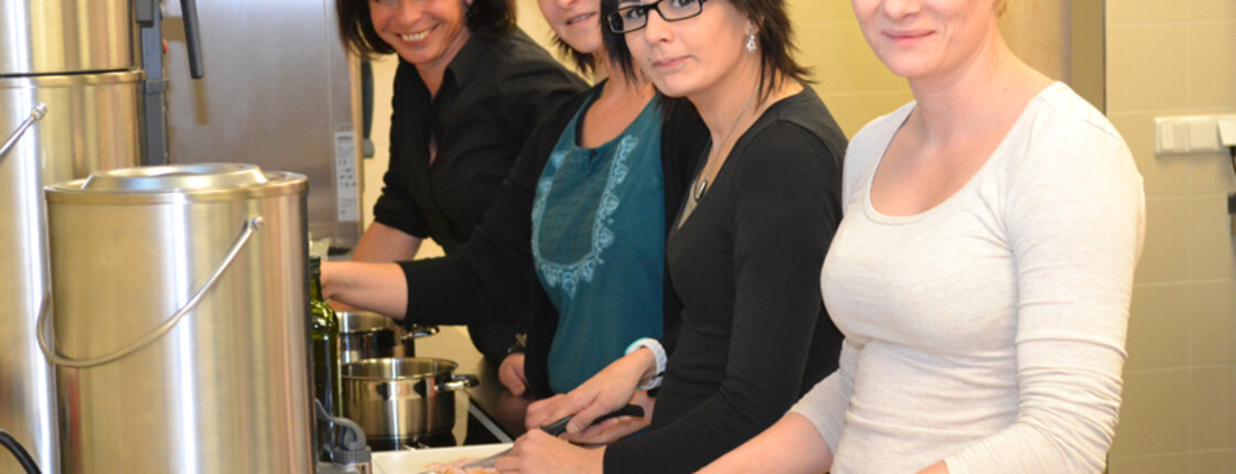 Vier Frauen mittleren Alters, die bis auf eine dunkle Haare haben, stehen in der Küche und schneiden Wurst auf. Sie schauen alle mit dem Kopf gebückt fröhlich in die Kamera. 