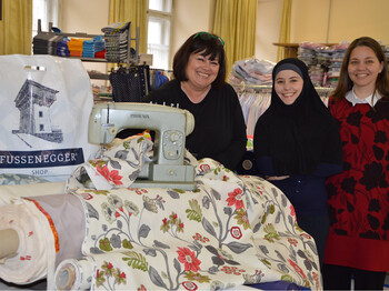 Zwei Frauen mittleren Alters stehen mit einem jungen, erwachsenen. weiblichen Füchtling in einem Nähgeschäft. Alle drei Frauen sind schlicht in Schwarz bzw. in Rot gekleidet. Im Vordergrund steht eine grüne Nähmaschine. Der Hintergrund ziert Ansammlungen von Stoffen auf Regalen. 