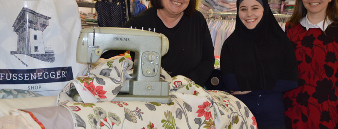 Zwei Frauen mittleren Alters stehen mit einem jungen, erwachsenen. weiblichen Füchtling in einem Nähgeschäft. Alle drei Frauen sind schlicht in Schwarz bzw. in Rot gekleidet. Im Vordergrund steht eine grüne Nähmaschine. Der Hintergrund ziert Ansammlungen von Stoffen auf Regalen. 