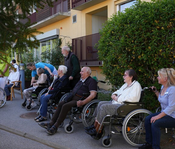 Mehrere Bewohnerinnen und Bewohner eines Pflegewohnhauses sitzen im Freien beisammen und verfolgen gemeinschaftlich das Geschehen rund um das festliche Maibaumaufstellen.