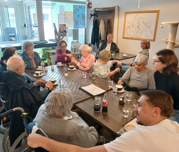 Gruppe von Bewohner*innen sitzt am Tisch im Katzencafé, trinkt Kaffee und tauscht sich während des Ausflugs aus