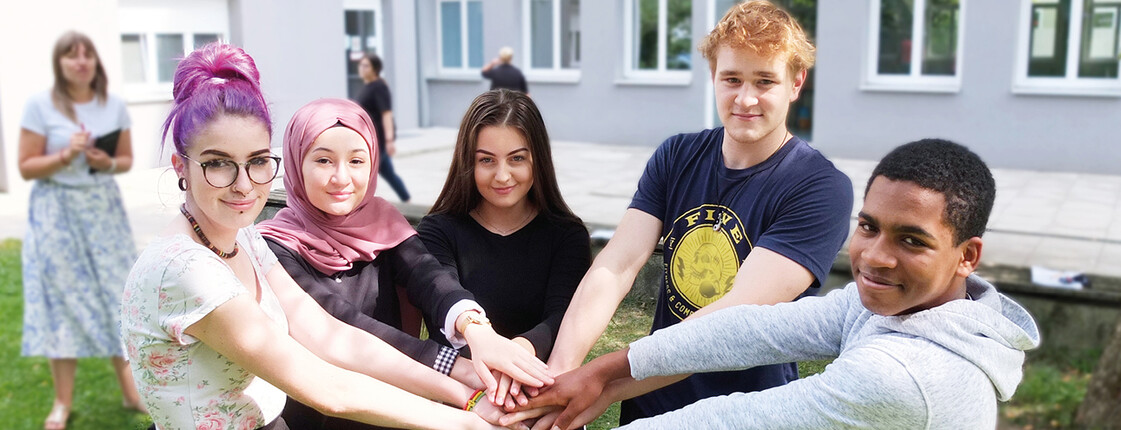Schülerinnen und Schüler der Caritas Schule Wiener Neustadt 3 Schülerinnen und 2 Schüler stehen im Halbkreis beieinander im Grünen im Hof der Schule und Halten die Hände übereinander. Im Hintergrund steht verschwommen eine Schülerin mit Buch in der Hand. Copyright Caritas Schule Wiener Neustadt