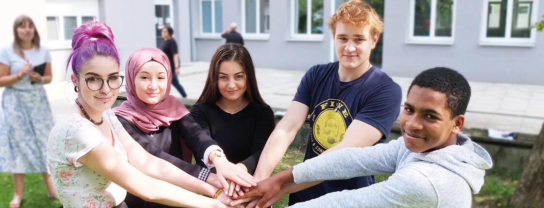 3 Schülerinnen und 2 Schüler stehen im Halbkreis beieinander im Grünen im Hof der Schule und Halten die Hände übereinander. Im Hintergrund steht verschwommen eine Schülerin mit Buch in der Hand. Copyright Caritas Schule Wiener Neustadt