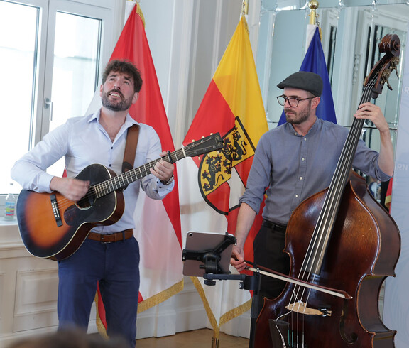 Zwei Musiker spielen Gitarre und Kontrabass vor Fahnen, im Raum eines Festakts