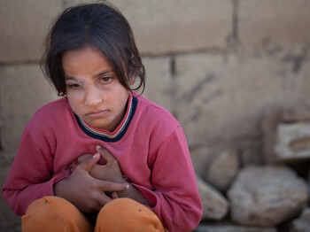 Kleines libanesches Mädchen sitzt mit verschränkten Händen vor einer mit Staub überzogenen Mauer. Sie trägt ein rotes Oberteil und eine orange Hose. Sie wirkt in sich gekehrt. 