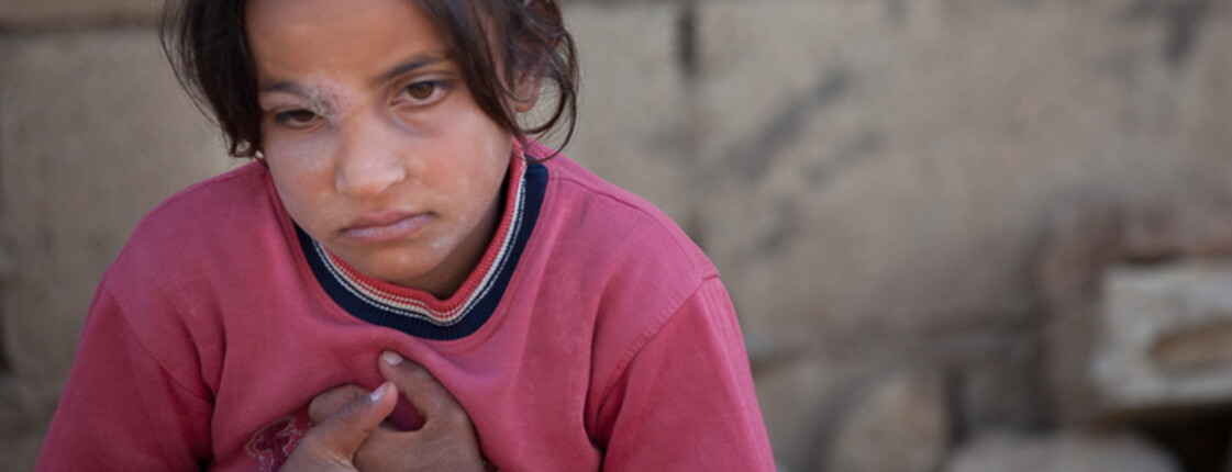 Kleines libanesches Mädchen sitzt mit verschränkten Händen vor einer mit Staub überzogenen Mauer. Sie trägt ein rotes Oberteil und eine orange Hose. Sie wirkt in sich gekehrt. 