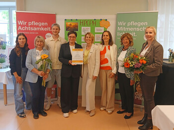 Gruppe aus Projektleitung, Pflege, Politik und Trägervertretungen steht mit Urkunde und Blumen vor HPCPH‑Tafel und Caritas‑Rollups