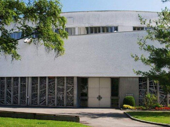 Außenansicht der Pfarre Bruder Klaus. Es ist ein größes, etwas in die Jahre gekommenes Gebäude, das grau angestrichen ist. 