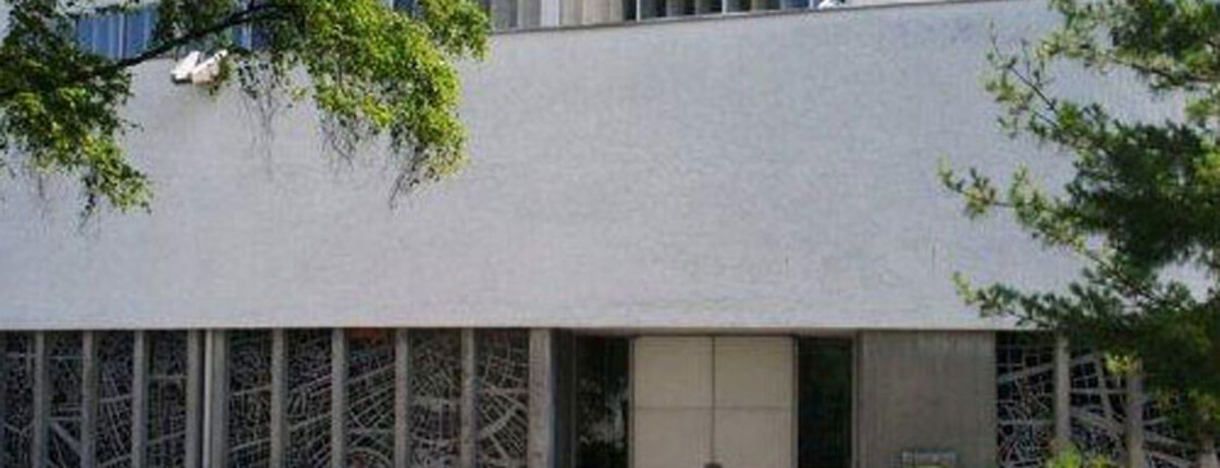 Außenansicht der Pfarre Bruder Klaus. Es ist ein größes, etwas in die Jahre gekommenes Gebäude, das grau angestrichen ist. 