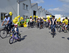 Ca. 20 Menschen mit an ihren Rädern gelb befestigten Luftballonen befinden sich auf einem großen asphaltierten Platz. 
