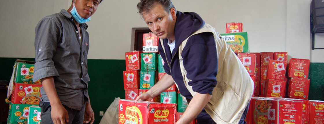 Zwei Männer unterschliedlicher Nationalitäten tragen Lebensmittelpakete ins Lager. Ein Mann hält gerade ein solches in der Hand. 