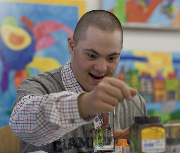 Mensch mit Behinderung widmet sich völlig dem Malen mit Aquarellfarben. Freude steht ihm im Gesicht. 