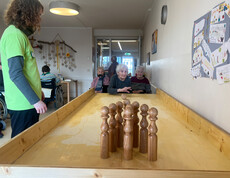 Bewohner*innen sitzen im Aufenthaltsraum und verfolgen das Tischkegeln auf der langen Holzbahn. 