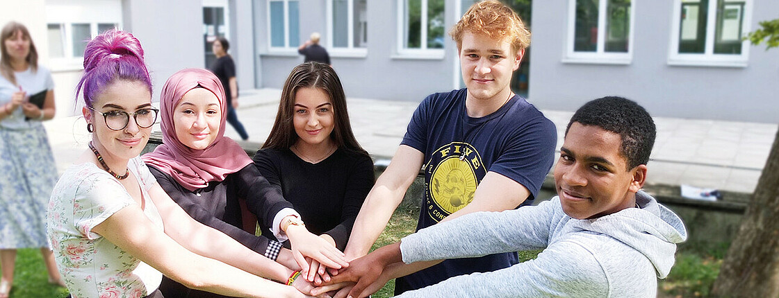 3 Schülerinnen und 2 Schüler stehen im Halbkreis beieinander im Grünen im Hof der Schule und Halten die Hände übereinander. Im Hintergrund steht verschwommen eine Schülerin mit Buch in der Hand. Copyright Caritas Schule Wiener Neustadt